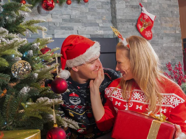 Grateful woman thanks her sweetheart for a surprise Christmas present. Cute couple celebrating festive December holidays in embrace of warm home next to beautifully decorated Christmas tree.