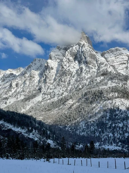 View from the valley to the majestic high mountain covered with fresh snow. White clouds gather around the snowy peak. Beautiful winter wonderland at remote high altitudes of wild Albanian Alps.