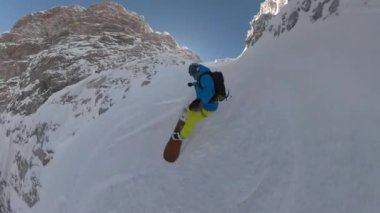 Male snowboarder freeriding down the steep mountain slope on fresh powder snow. Young man spraying snow while snowboarding freshly snow-covered terrain in the embrace of picturesque high mountains.