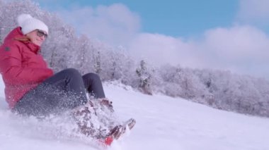 SLOW MOTION: Young lady laughing while riding sled on fresh snow on a sunny day. Cheerful woman in warm outfit spraying snow while sledding downhill on gorgeous sunny winter day after fresh snowfall.
