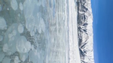 VERTICAL, CLOSE UP, DOF: Strong gusts of wind blow across the scenic frozen Lake Abraham on a sunny winter day. Stunning shot of Canadian Rockies behind a glassy frozen lake full of methane bubbles.