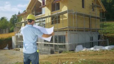 SLOW MOTION, COPY SPACE, CLOSE UP: Construction site manager analyses floor plans while also looking at house. Contractor looks at blueprints while standing outside a modern house under construction.