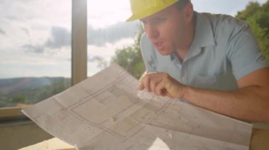 SLOW MOTION LENS FLARE CLOSE UP: Young builder wearing a yellow hard hat blows sawdust off blueprints lying on his workbench. Contractor inspects floor plans in a large sunlit room under construction