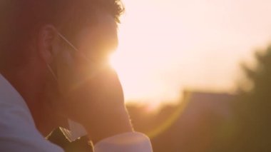 Yavaş Hareket, Yaklaş, El HELD, LENS FLARE, Dof: Genç beyaz adam altın gün batımını izlerken cerrahi maskesini çıkarıyor. İnsan gün doğumunu izlerken maskesini çıkardığı için rahatlar.