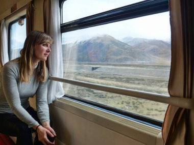 CLOSE UP Young woman rides along Trans-Himalayan Railway and observes the breathtaking landscape. Female traveler daydreams while looking through the window of train travelling across the Himalayas. clipart