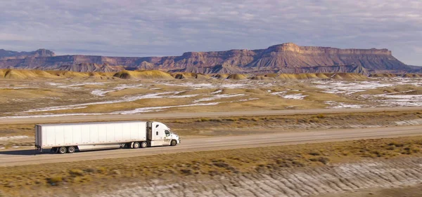 stock image AERIAL: Semi-trailer truck speeds along the motorway running across the desert in Utah. Wintry desert landscape surrounds truck driving down the interstate highway running across rugged US wilderness.