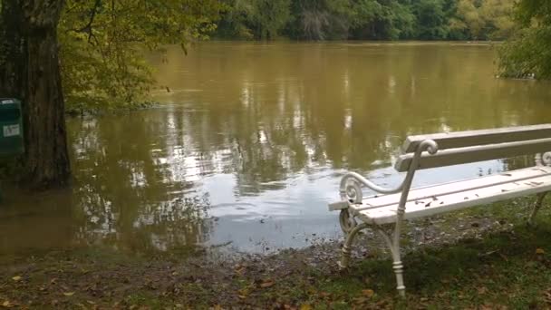 秋雨过后 白色的公园长椅在一条溢出的河流的脏水里 浑浊的洪水淹没了河岸和满溢的草坪和树木 大雨倾盆后严重水浸 — 图库视频影像