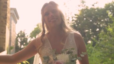 Neşeli gelin, altın güneş ışığıyla bir ağaca yaslanmış, neşe saçıyor. Zarif, beyaz gelinlikli, doğal makyajlı ve gün batımında gür yeşil doğada dalgalı saçlı güzel bir bayan.