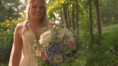 Mutlu gelin, altın ışıkta kameraya doğru buket tutarak yaklaş. Muhteşem yaz günbatımında düğün gününde pastel renkte güzel bir buket çiçek sergiliyor.