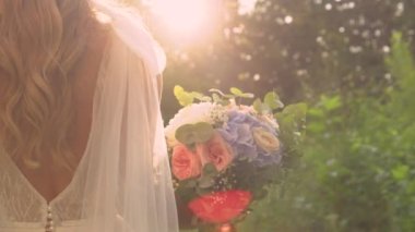 LENS FLARE, Altın ışığı kapatın ve gelin muhteşem bir gelin buketiyle yürüyor. Pastel tonlarında yaz çiçekleriyle muhteşem bir düğün buketi ve altın gün batımında beyaz elbiseli güzel bir gelin..