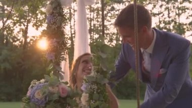 Yakışıklı damat, salıncakta sallanan yeni evli karısını öpüyor. Gülümseyen yeni evliler, düğün günlerinde altın gün batımında romantik bir an yaşarken birbirlerine şefkat gösterirler..