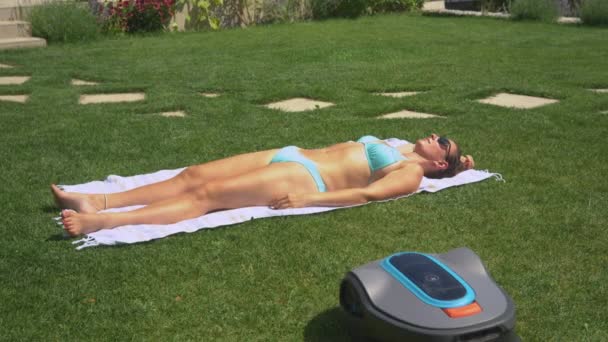 年轻女人在日光浴时被机器人割草机撞倒会做出反应 在阳光明媚的夏日 女士正在晒日光浴 而自动割草机则在自家后院修剪绿草 — 图库视频影像