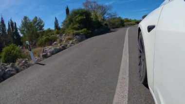 Adriyatik manzarası boyunca asfalt yolda giden bir araba var. Akdeniz büyümesiyle çevrili dolambaçlı yolun ortasındaki düz beyaz çizgiden geçen ön tekerleğin görüntüsü