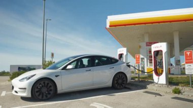 TESLA SUPERCHARGER, ZAGREB, CROATIA, 05.05.2023: Tesla şarj noktası park halindeki elektrikli araba pilini dolduruyor. Beyaz renkli BEV, Supercharger 'da konnektörle hızlı şarj ediliyor.