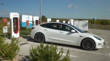 TESLA SUPERCHARGER, ZAGREB, CROATIA, 05.05.2023: Tesla şarj noktası park halindeki elektrikli araba pilini dolduruyor. Beyaz renkli BEV, Supercharger 'da konnektörle hızlı şarj ediliyor.