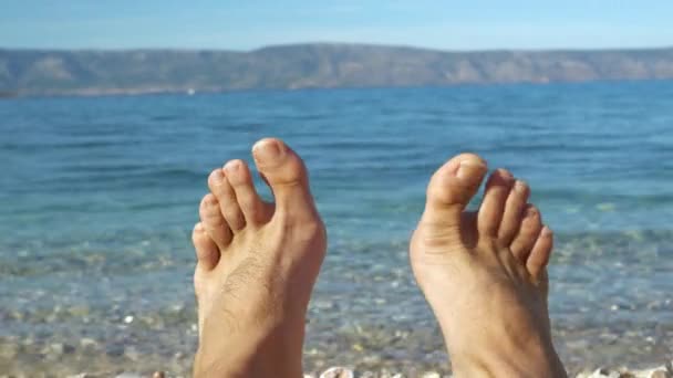 在蓝色海边阳光灿烂的亚得里亚海海滩上赤脚摆动 年轻人在放暑假 他正坐在卵石般的海岸上 望着晶莹如茵的大海翻滚而过 — 图库视频影像