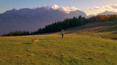 Genç bir kadın köpeğiyle çayırlık bir tepede koşarken nefessiz kalıyor. Açık havada spor aktiviteleri için güzel bir sonbahar günü. Güzel gün batımı ışığı manzaralı dağlık arazide parlar..
