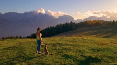 Çekici kadın, köpeğiyle birlikte bir tepenin üstündeki dağların manzarasına hayran. İnanılmaz güneşli bir sonbahar günü. Açık hava eğlencesi için. Resimli dağların kucaklaması ve evcil hayvanlarla kaynaşma zamanı..