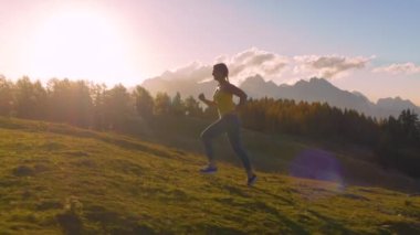 Yaklaş, LENS FlaRE: spor giyimli genç bayan çimenli bir sırt üzerinde koşar. Altın sonbahar güneşinin ilk ışınları Alp arazisine dökülmeye başlarken, o, pitoresk bir patika boyunca koşuyor..