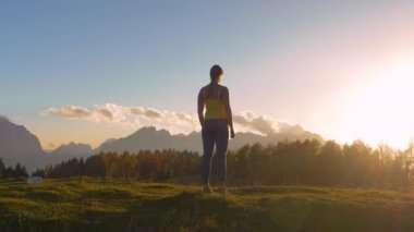 Yaklaş, LENS FlaRE: spor kadın güzel gün batımını takdir etmek için tepede durdu. Tepeye kadar uzanan altın ışıkta yüksek dağları görerek kendini ödüllendiriyor..