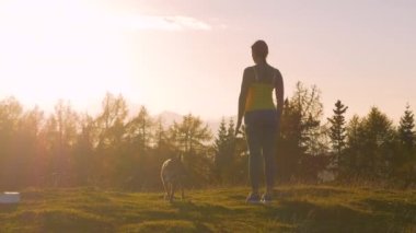 Yaklaş, LENS FlaRE: bir köpek ile sportif kadın çimenli zirvede gün batımına hayran. Alp dağlarının üzerinde parlayan altın sonbahar güneşinin son ışıklarını görmek için kendini durdurdu..