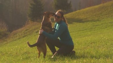 Gülümseyen genç bir kadın, sevimli kahverengi köpeğine kuyruğunu sallayarak sarılıyor. Altın sonbahar günbatımında güzel yeşil bir çayırda yapılan başarılı geri çağırma eğitiminden sonra köpeği okşuyor..