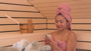 Saunada otururken akıllı telefondan mesaj atan gülümseyen Asyalı kadın. Rahatlatıcı ve rahatlatıcı bir sauna tedavisi yerine cep telefonu ve sosyal medya ile dikkati dağılıyor..