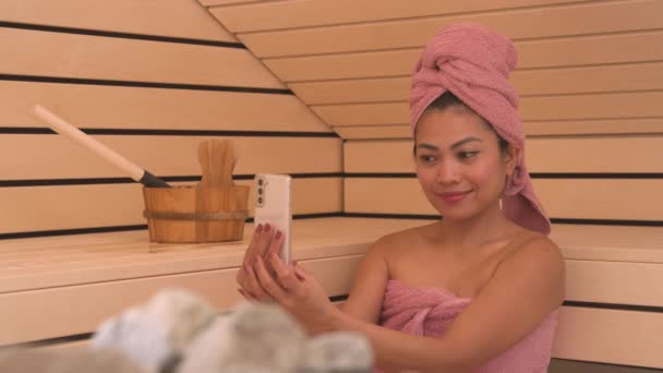 漂亮的菲律宾女人在木制芬兰桑拿用电话自慰 她没有享受轻松舒适的桑拿待遇 而是被手机和社交媒体弄得心烦意乱 — 图库视频影像