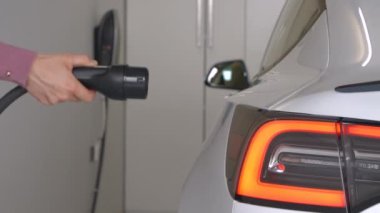 Kadın garajda motor aküsünü şarj etmek için beyaz bir BEV takıyor. Elektrikli aracı kullandıktan sonra siyah güç kablosunu bağlıyor. Elektrikli arabayı şarj etmek için zahmetsiz bir sistem.