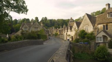 Köprüden geçerek taşra köyüne doğru büyüleyici taş evlerle. Yeşil bir tepenin altında asfalt bir yol boyunca yerleştirilmiş güzel Castle Combe 'da resimli teraslı evler.
