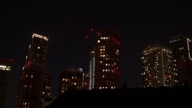 Akşam saatlerinde iş yerleri ve konut binalarıyla dolu güzel bir şehir manzarası. Londra bölgesindeki yüksek gökdelenlerin camlarında gece karanlığında ışıklar parlıyor..