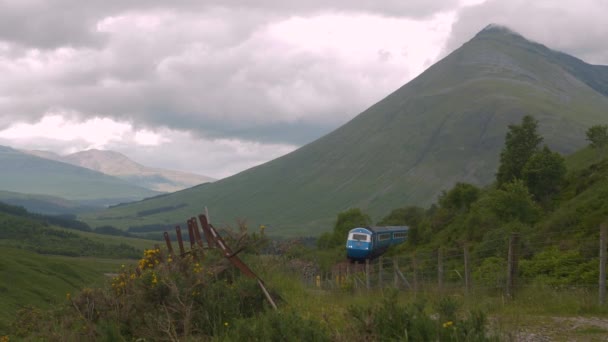 曇った夏の日にカーブを通過する青い旅客列車で美しいスコットランドの風景 広大な緑の谷を経由する絵画的な鉄道ルート それらの上に草の多い山々 — ストック動画
