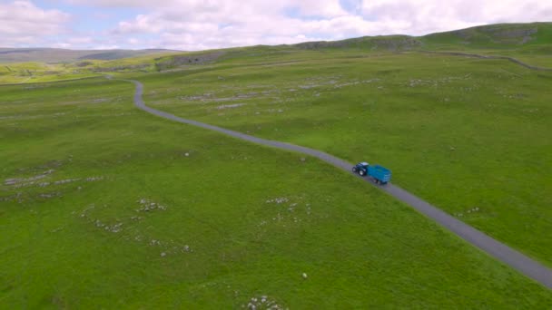 Aerial ブルートラクターは 新鮮なカットされた草でいっぱいのトレーラーで巻く単一のトラックの国道に沿って運転します ヨークシャー デールズ国立公園の石で囲まれた牧草地の田園地帯を転がす — ストック動画