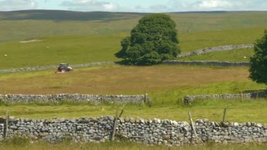 Kuru taş duvarlarla çevrili yeşil çayır ve yem için yaz çimlerini biçen bir traktör. Çiftçilik için modern ekipman kullanımı. Yorkshire Dales Ulusal Parkı 'nın kırsalında güneşli güzel bir gün.