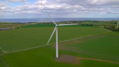 Rüzgarlı İskoç sahillerindeki yeşil tarlaların ortasında dönen yel değirmenleri. Sürdürülebilir elektrik üretimi için doğal elementlerden yararlanan uzun ve görkemli modern yapılar.