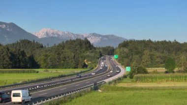 Ormanlarla çevrili güneşli Slovenya otoyolunda yakınlaşan arabaların uçan bir videosu. Arka planda övünerek dağlara doğru Gorenjska otoyolunda yaz yolculuğu.