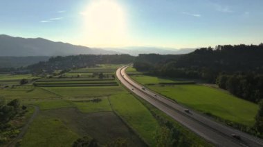Güneşli otoyolun, yaz aylarında giden arabalarla çekilmiş görüntüsü var. Bereketli yeşil kırsal alanlar ve güneş ışığında gelişen ekinler. Arka planda bir tepeye tünemiş güzel bir köy