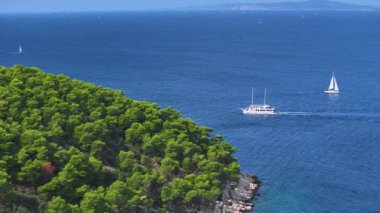 Hvar 'ın yemyeşil çam ağacı boyunca seyreden iki teknenin manzaralı görüntüsü. Yelkenli ve yat, Adriyatik 'in yemyeşil kıyı şeridi yakınlarındaki derin mavi denizlerini keşfederken buluşuyor..