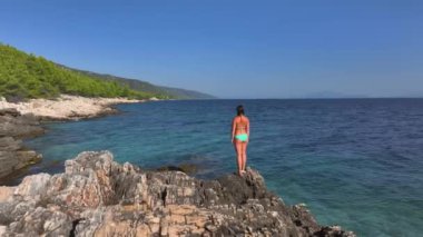 Bikinili tanınmayan kadın kayalık çıkıntıda duruyor ve Hvar 'ın kayalık kıyılarını gözlemliyor. Hırvatistan 'ın Dalmaçya kentindeki gözlerden uzak muhteşem plaja bakan genç bir bayan turistin sahne çekimi