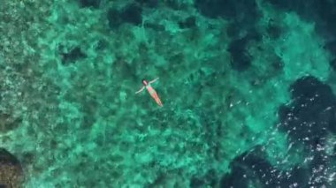 Mavi bir bikini giymiş, açık mavi bir denizde huzur içinde yüzen genç bir kadın. Kolları, rahatlatıcı bir meditasyonda gerilmiş. Dalgalar tarafından nazikçe sallanıyor.