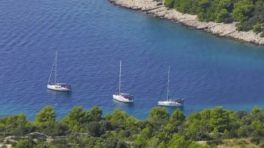 Üç lüks yelkenli engebeli bir Akdeniz kıyısının yanında demirlemiş kaygısız yaz tatilinin simgesi. Hırvatistan 'ın Hvar adasının manzaralı kıyılarına demirlemiş yelkenliler