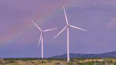 Rüzgar çiftliği, yenilenebilir enerji teknolojisini sergileyerek gökyüzüne karşı dimdik ayakta duruyor. Rüzgâr türbinlerinin üzerinde, sürdürülebilir enerjiyi sembolize eden, engebeli bir araziye kurulmuş çarpıcı bir gökkuşağı kavisleri..