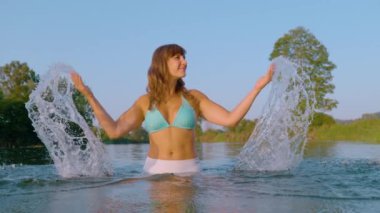 Bikinili çekici genç bir kadın kollarını kaldırıp zarif bir şekilde su sıçratırken nehirde duruyor. Sıcak yaz günlerinde el değmemiş ve ferahlatıcı nehirde yıkanmayı sever..