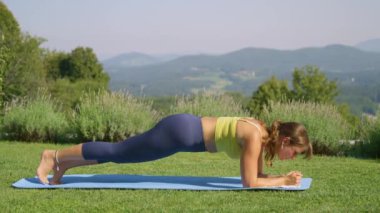 Çekirdek dayanıklılığını artırmak için bacağıyla yorucu tahta pozu veren formda bir kadın. Aktivite kıyafetli sporcu kadın bahçedeki minderinin üzerinde gelişmiş bir yoga gösterisi yapıyor..