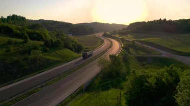 AERIAL, LENS FLARE: Calm morning and flowing traffic on the winding highway leading through a green landscape. A beautiful and scenic drive past lush forests and hilly countryside in golden light.