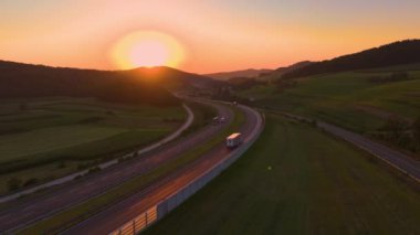 AERIAL, LENS FLARE: Meandering motorway glows in orange hue of setting summer sun. Highway with smooth flowing traffic winds past green fields and lush forests stretching across the hilly countryside.