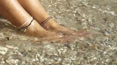 CLOSE UP: Sunlit feet adorned with anklets, caressed by gentle seaside waves over a pebbled beach. Serene moment of summer relaxation and refreshment at a beautiful Adriatic beach in sunny Dalmatia.