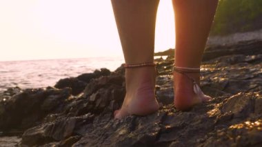 Çıplak ayaklı genç bayan, batan güneşin altın ışınlarının engebeli kayaları yansıttığı bir deniz kıyısında duruyor. Ayak bilekleri basit ve güzel deniz kabuğu bilezikleriyle süslenmiş..