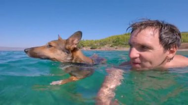 Köpeğiyle yüzen gülümseyen genç bir adamın güzel bir yaz anı. Neşeli adam sıcak ve güneşli bir günde ferahlatıcı denizde sevimli bir köpekle birlikte yüzer. Hırvat deniz kıyısında aktif tatil.