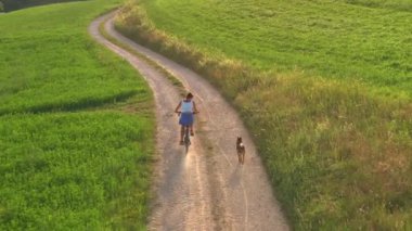 Güzel yeşil çayırlar arasında çakıllı bir köy yolunda köpeğiyle bisiklete binen sportif bir kadın. Açık hava aktivitelerinin keyfi ve kırsal alanda sevilen bir evcil hayvanın arkadaşlığı..